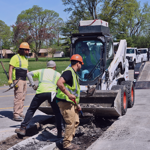 Public Works crew repairing pavement with a skid steer and hand tools in a parking lot